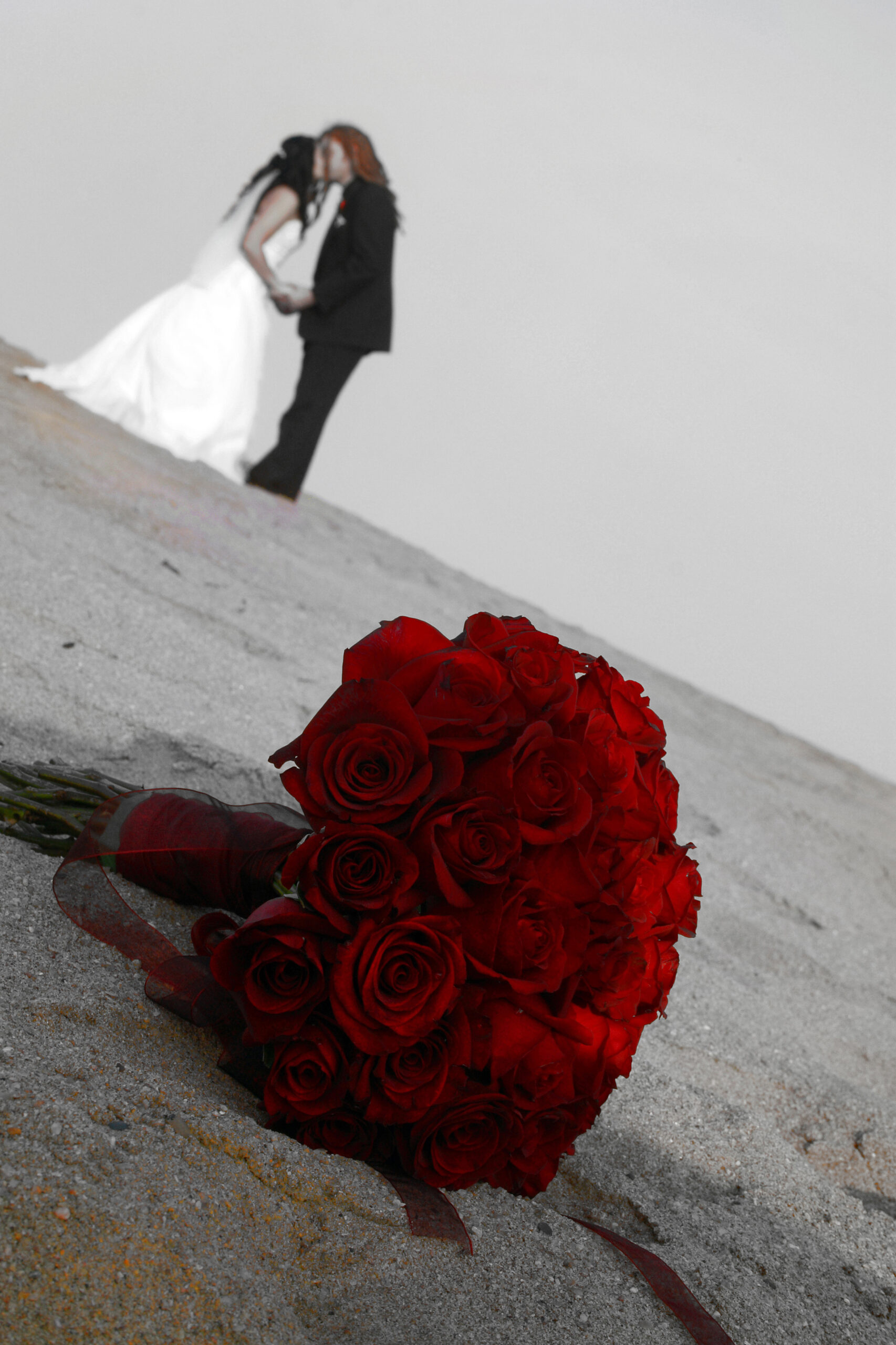 Selektiv koloriertes Foto eines küssenden Brautpaares mit einem Bouquet roter Rosen im Vordergrund