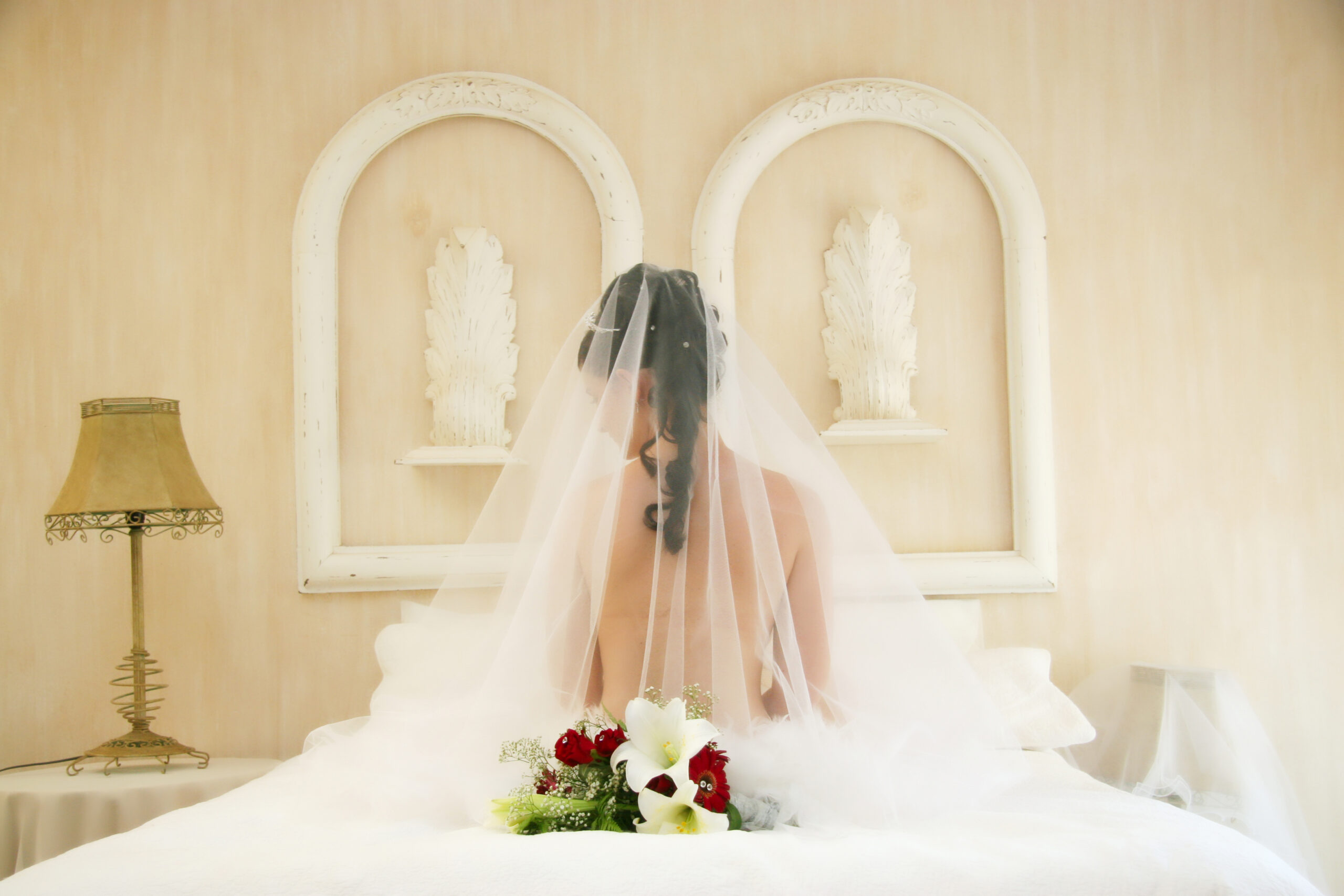 Braut in weißem Kleid und Schleier sitzt vor gotischen Fenstern und hält einen Brautstrauß