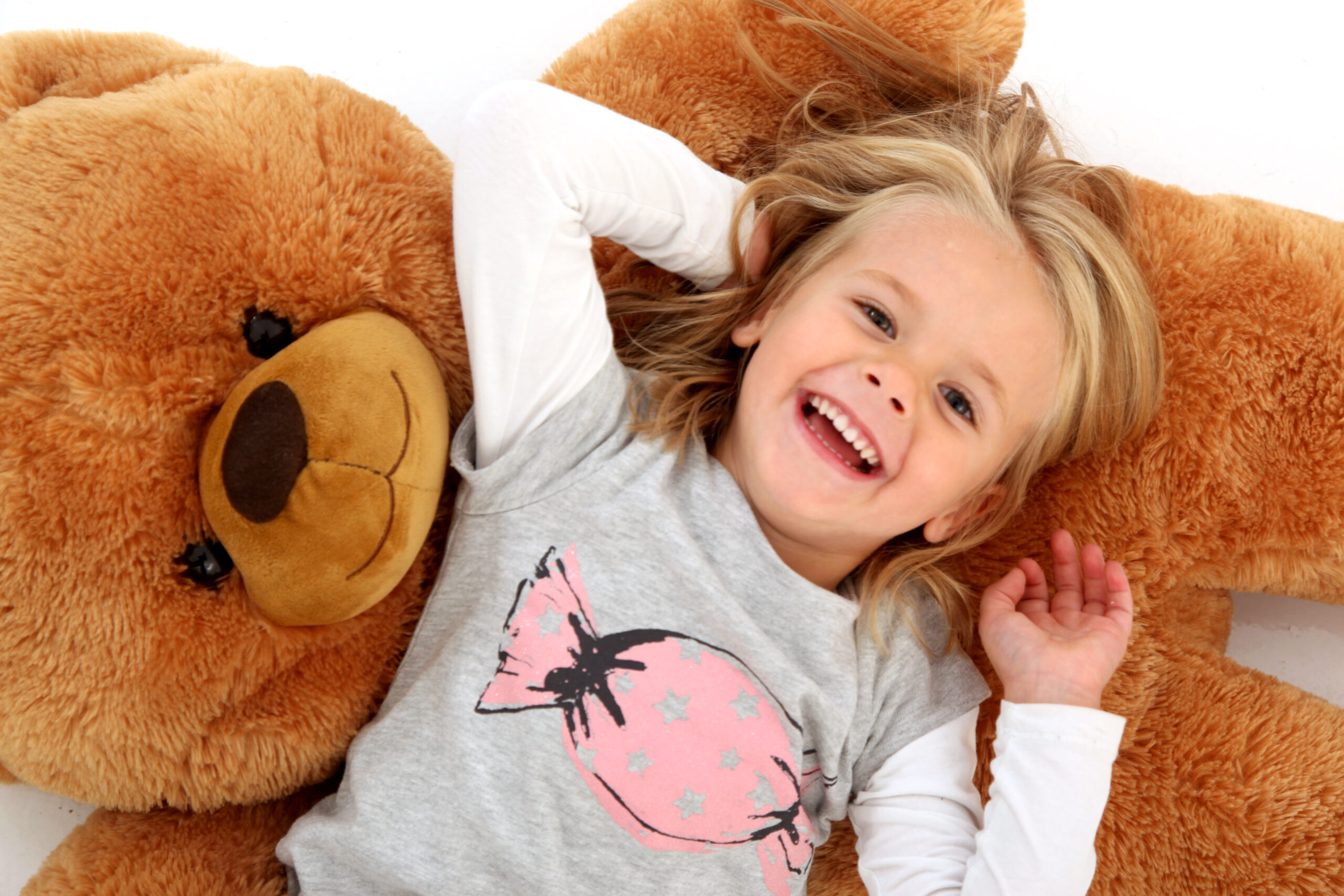 Ein lächelndes junges Mädchen liegt auf dem Rücken und umarmt einen großen, flauschigen Teddybären.