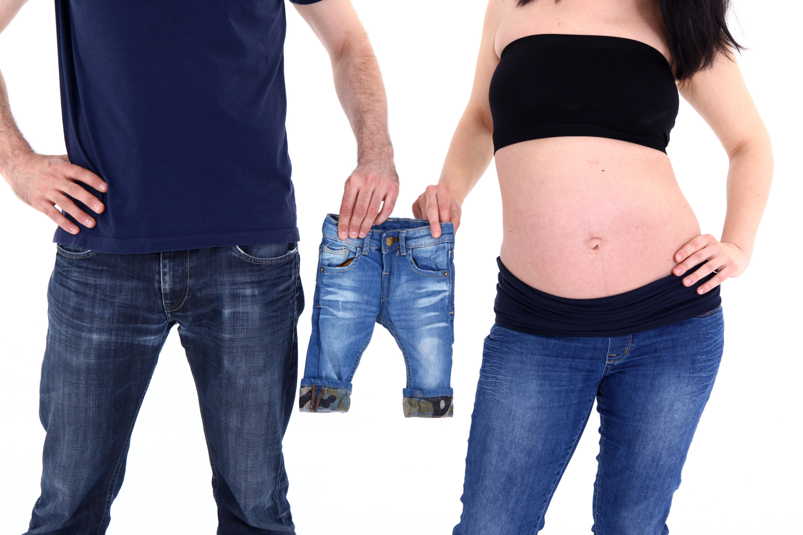 Ein Mann und eine schwangere Frau stehen nebeneinander, er hält eine kleine Jeanshose und sie umfasst ihren Babybauch.