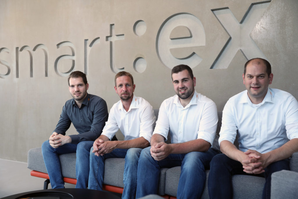 Vier lächelnde Männer in Hemden sitzen auf einer Couch vor einer Wand mit dem Wort 'smartex'.