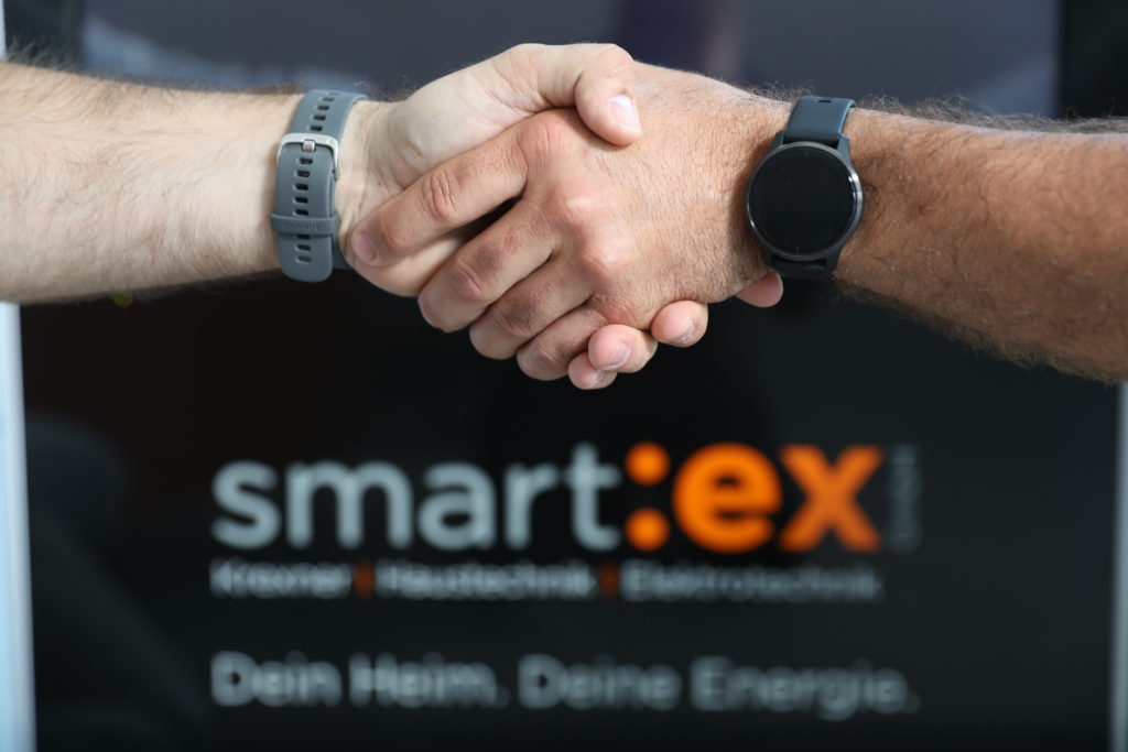 Handschlag mit Armband und Smartwatch vor einem Plakat mit der Aufschrift 'smart:ex'