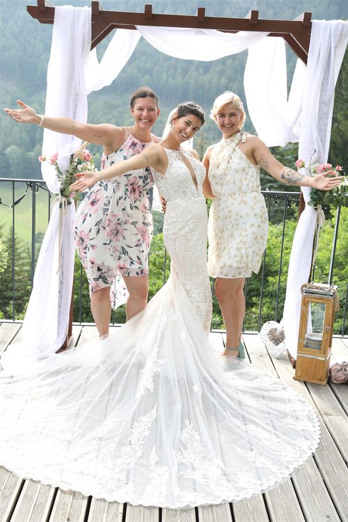 Braut in weißem Kleid posiert mit zwei Freundinnen in Sommerkleidern im Freien