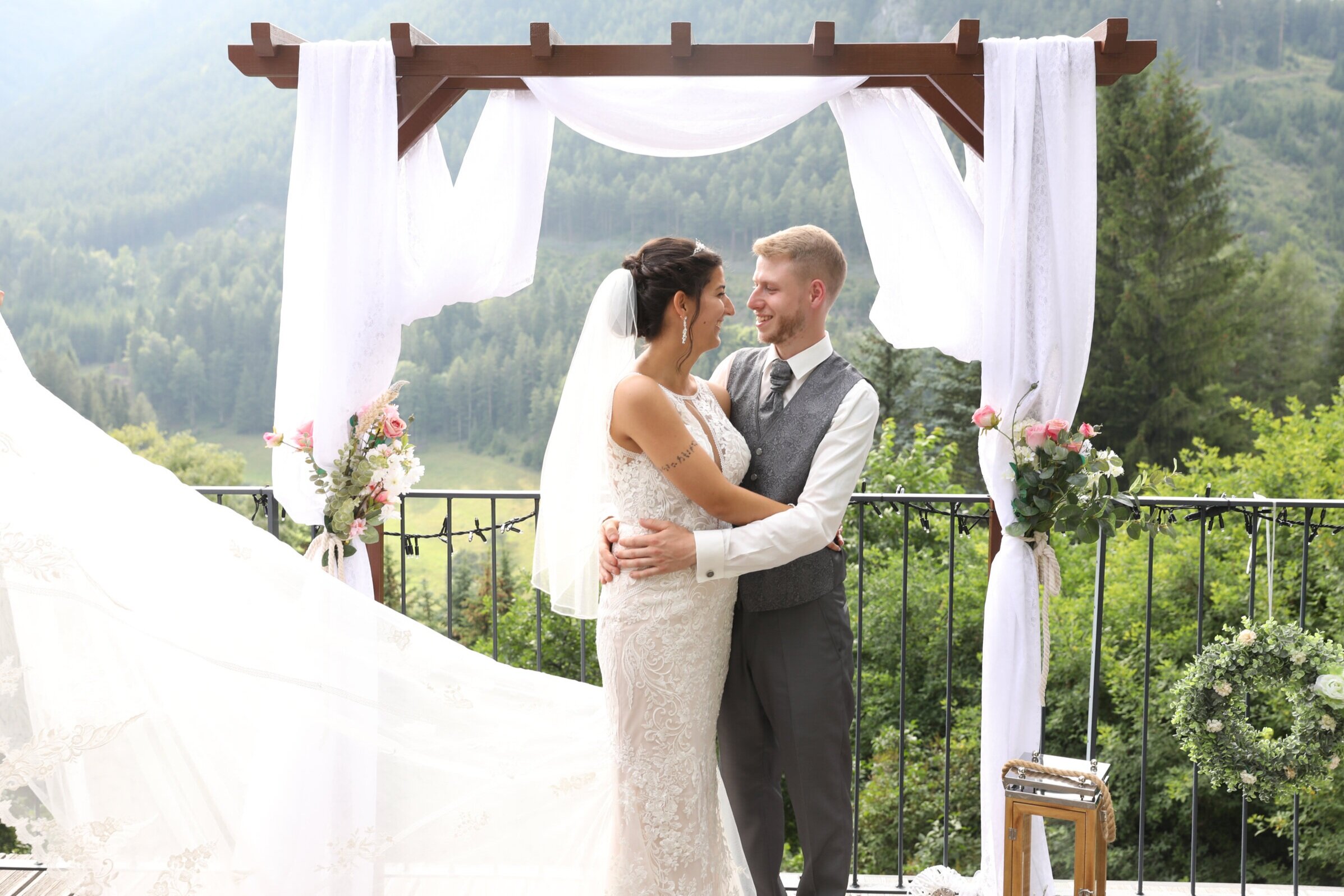 Brautpaar umarmt sich unter einem dekorierten Hochzeitsbogen mit Bergblick im Hintergrund
